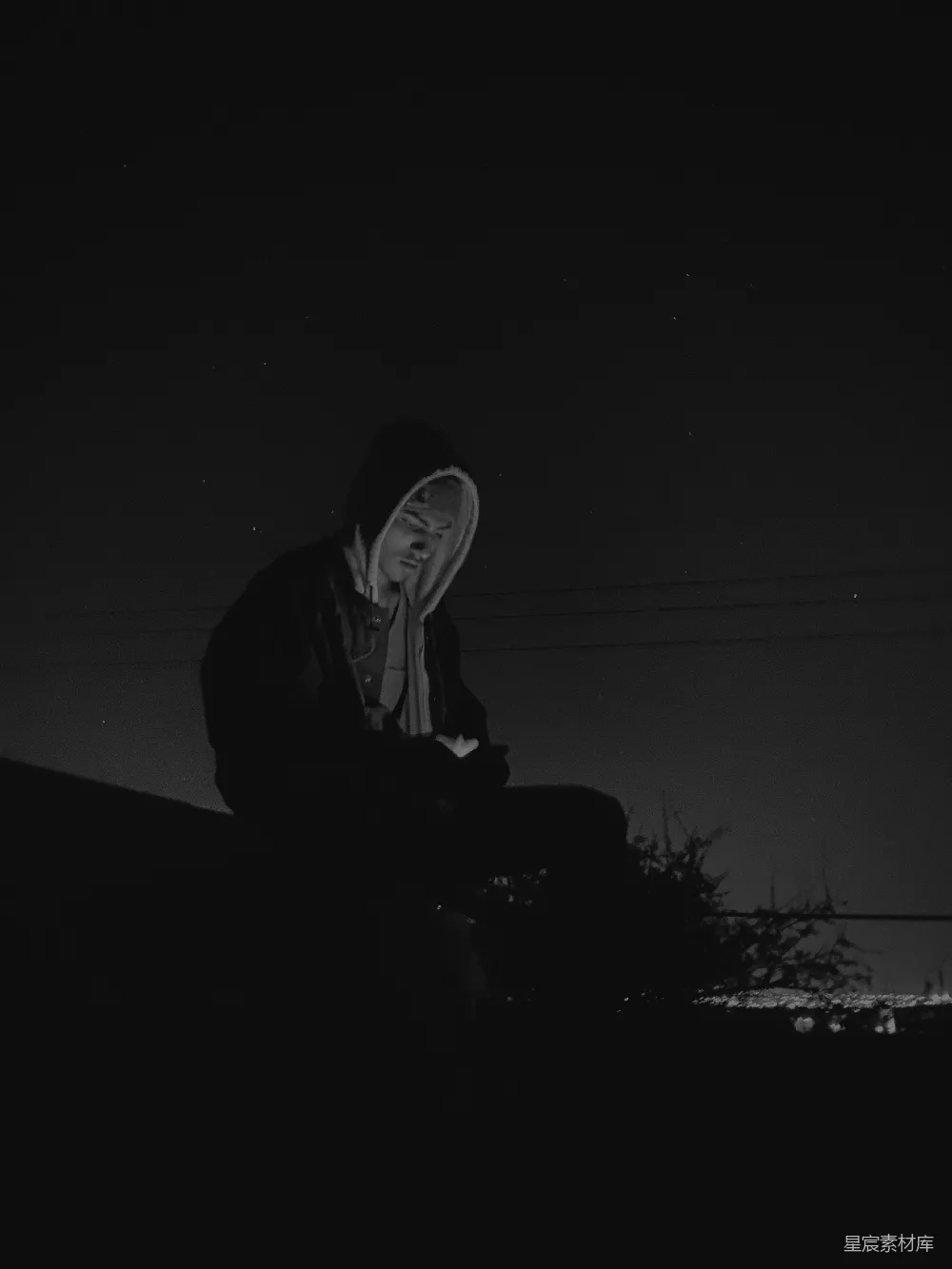 这张黑白照片中，一个穿着连帽衫的人在夜晚的环境中独坐，背景是昏暗的天空和电线。他似乎在沉思，画面充满了宁静与孤独的氛围，适合用作情感故事或独立思考的背景图。