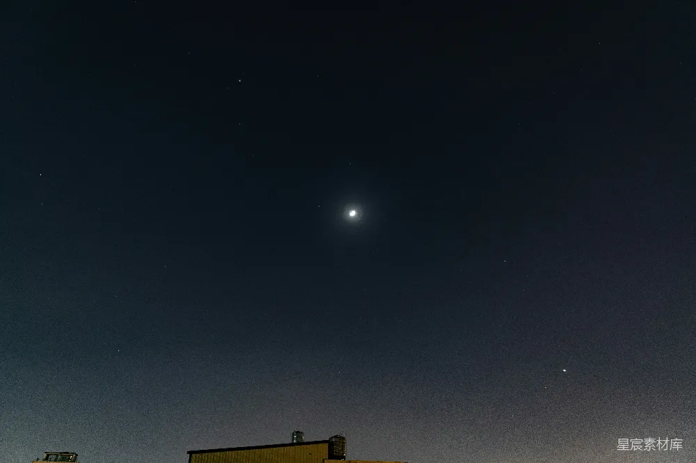 这张图片展示了一个宁静的夜空场景，明亮的月亮位于画面中央，周围点缀着无数璀璨的星星。底部可以看到一些建筑物的屋顶，增添了画面的深度。整体色调偏暗，突出了夜空的神秘与美丽。适合用于展示自然风光、宁静氛围的背景或作为摄影爱好者的素材。