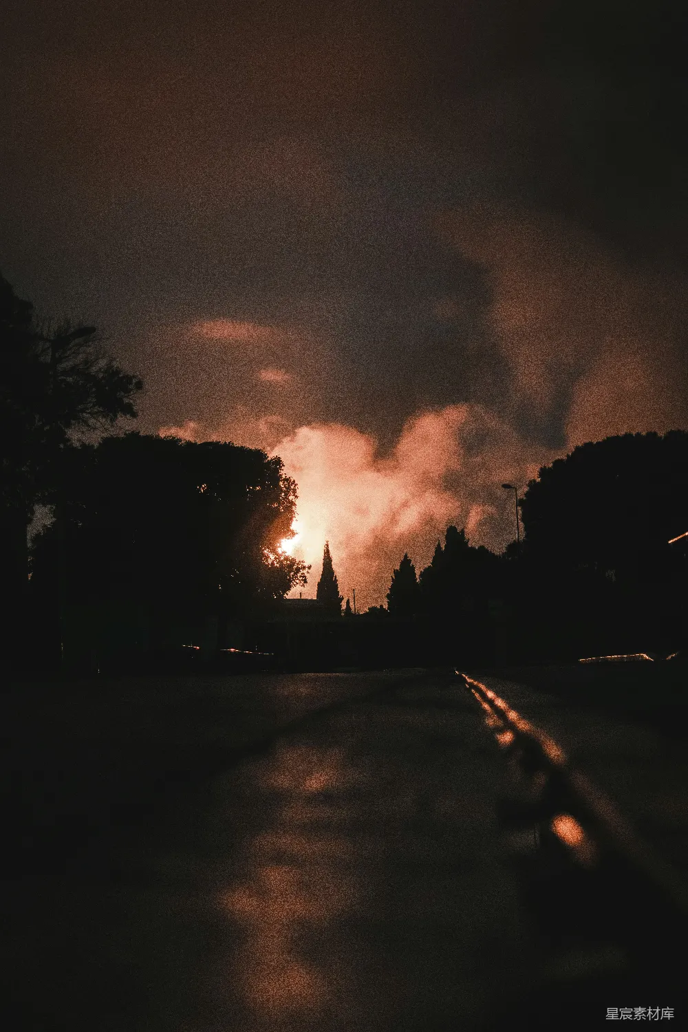 这张图片展示了一个黄昏时分的道路景象，太阳的余晖透过云层洒在地面上，形成了美丽的光影效果。路的两侧是树木的剪影，远处的建筑物在夕阳的照射下显得朦胧而宁静。整体画面充满了自然与宁静的氛围，适合用作旅行、自然或宁静生活的背景图片。