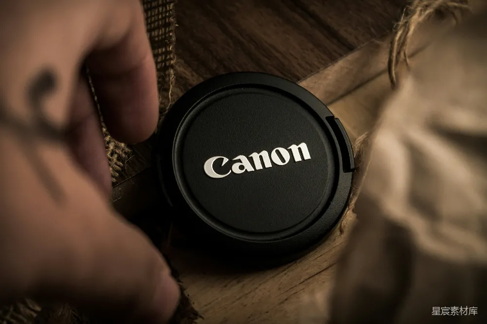图片展示了一只手拿着Canon镜头盖的特写镜头。镜头盖放置在木质表面上，背景模糊，突出了Canon的标志。这种镜头盖在摄影爱好者和专业人士中非常常见，用于保护镜头免受尘埃和划痕。