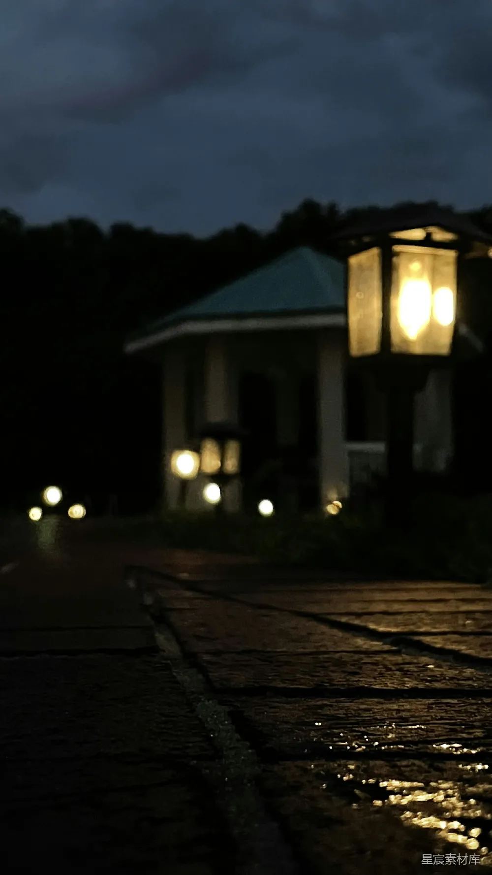 这张图片展示了一个宁静的夜晚，石板路上反射着路灯的温暖光芒。远处是一座乡村建筑，周围点缀着几盏柔和的灯光，营造出一种平和而温馨的氛围。适合用作夜景摄影、乡村生活或宁静氛围的背景素材。