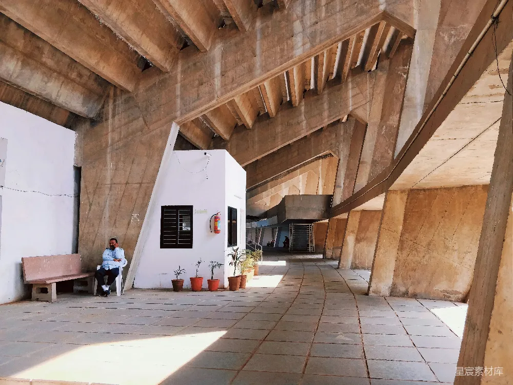 这张图片展示了现代建筑内部的一个宁静角落，宽敞的空间内，简洁的几何结构和自然光线交织，营造出一种宁静而富有艺术感的氛围。墙角处，一位男士正坐在长椅上，似乎在享受这份静谧。