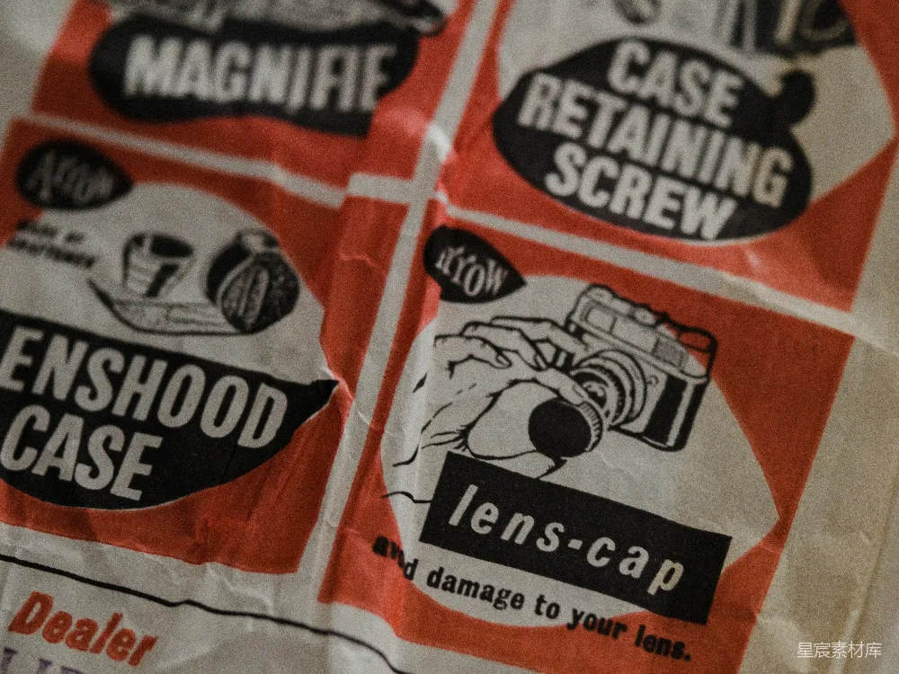 这张图片展示了复古相机包装纸的特写，纸张上印有“ENSFOOD CASE”和“lens-cap”的字样，以及一个手持相机的插图。包装纸的颜色主要是红色和白色，上面有黑色的印刷文字和图案。背景略显模糊，突出了包装纸的细节。这种图片适合用于复古