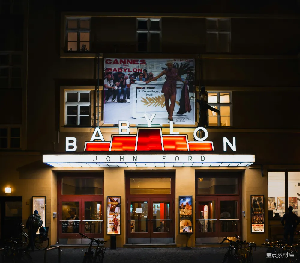 图片展示了柏林巴比伦剧院的夜景。剧院的招牌上用大字写着'BABYLON'和'JOHN FORD'，招牌下方的窗户透出温暖的灯光。剧院上方的广告牌上展示了戛纳电影节的海报，画面中有一位模特在走秀。剧院前的街道上有几辆自行车，夜晚的灯光与剧院的