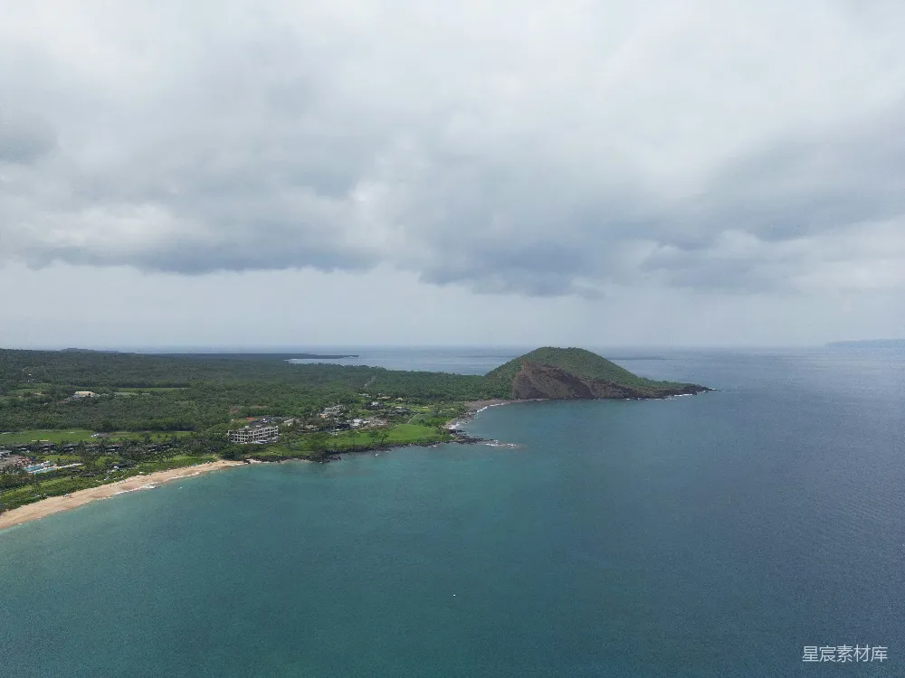 这张图片展示了海岸线旁的一座火山岛，周围被碧蓝的海水环绕。远处的岛屿上覆盖着茂密的绿色植被，近处的海滩上散布着一些建筑物。天空中飘着厚重的云层，为整个场景增添了一份宁静与神秘。适合用于旅游宣传或自然风光展示。
