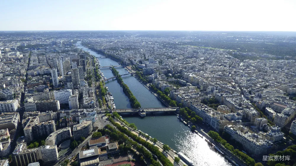 这张图片展示了巴黎的塞纳河及其周边的城市景观。从空中俯瞰，可以看到河流蜿蜒穿过密集的建筑群，桥梁连接两岸。阳光洒在河面上，反射出耀眼的光芒，周围环绕着郁郁葱葱的树木。这幅画面展示了巴黎这座城市的独特魅力与自然美景的和谐共存。