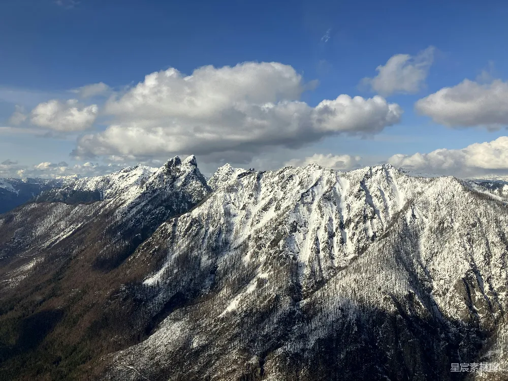 这张图片展示了连绵起伏的雪山，山顶覆盖着厚厚的积雪，云朵在蓝天中悠闲飘浮，呈现出一幅宁静而壮丽的自然景象。适合用于宣传冬季旅游、户外探险主题或自然风光的展示。