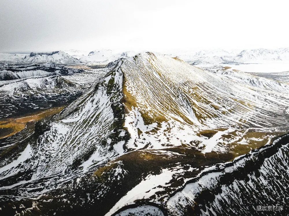 这张图片展示了雪山之巅的壮丽景色，白雪覆盖的山峰与金黄色的植被形成鲜明对比，远处的山脉和天空则为这幅画面增添了深度和广度。适合用于自然风光、冬季旅行或户外探险的宣传。