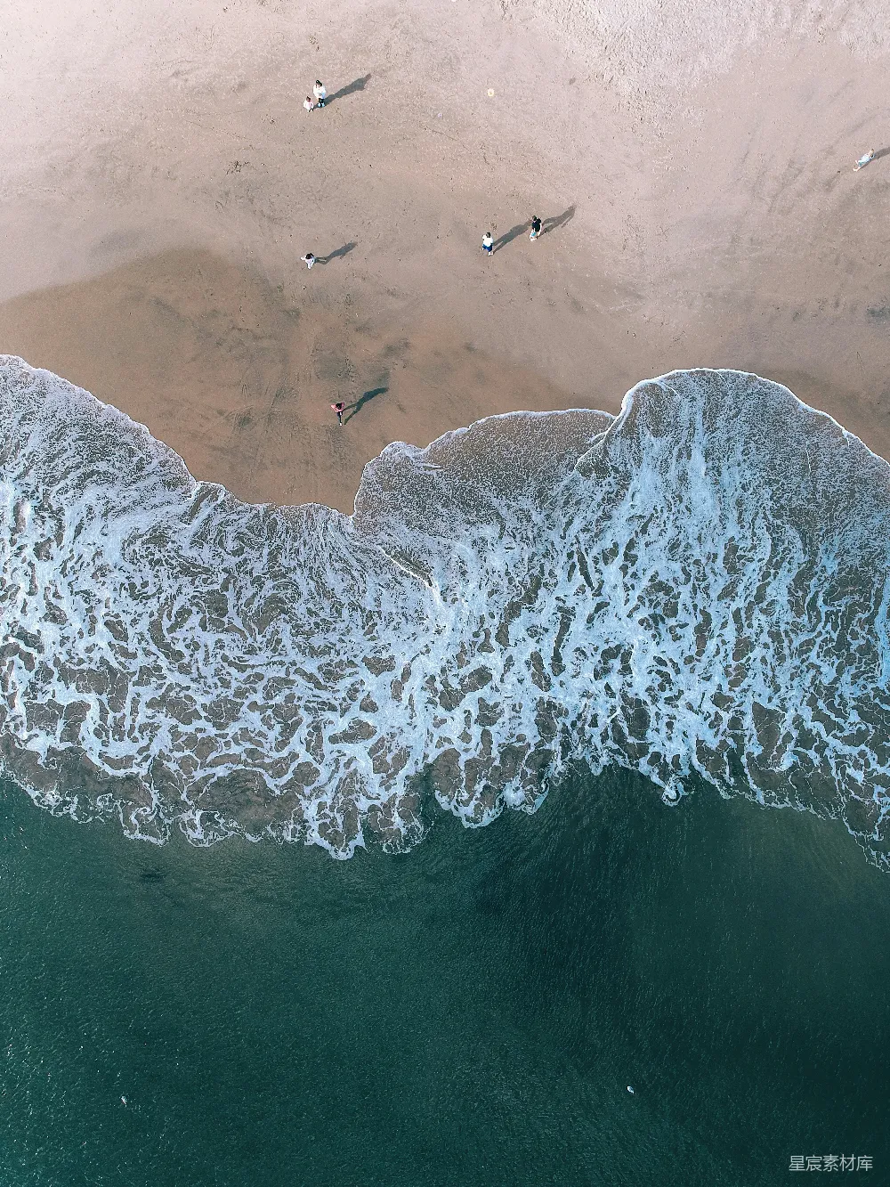 这张图片展示了从高空俯瞰海滩的视角，细腻的海浪轻轻拍打着沙滩，形成白色的浪花。沙滩上散布着几个休闲的人，享受着宁静的海岸风光。整体画面色调清新，给人一种宁静与放松的感觉，适合用于旅游、休闲或自然风光的展示。