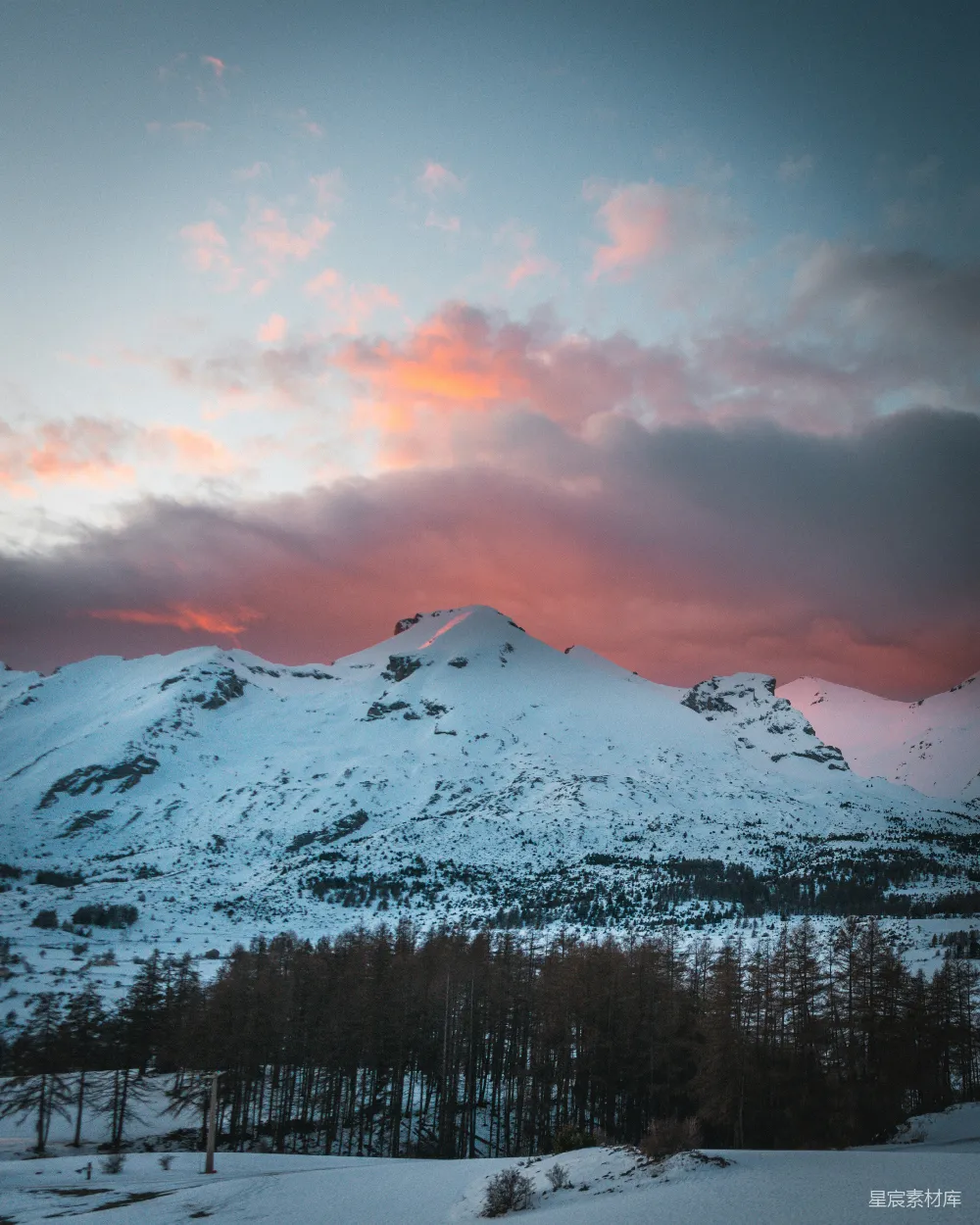 图片展示了一座被皑皑白雪覆盖的山峰，远处的天空被夕阳染成了粉红色，营造出一种宁静而美丽的冬日景象。前景是被雪覆盖的树木，远处的山峦在夕阳下显得格外壮丽。这幅画面适合用于自然风光、冬季旅行、摄影艺术等场景。