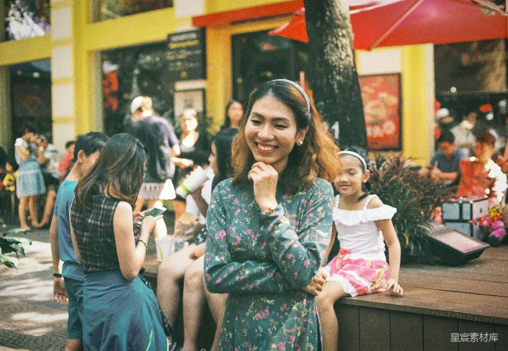 这张图片捕捉到了城市街道上的一幕温馨场景。一位母亲和女儿正坐在一处休息区，母亲微笑着，似乎在与女儿交谈。周围的环境中充满了生活气息，人们在背景中忙碌着，商店和树木点缀其间，营造出一种热闹而宁静的氛围。