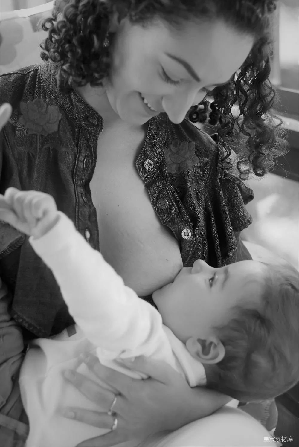 这张黑白照片捕捉了母乳喂养的温馨时刻。母亲温柔地抱着孩子，专注地看着孩子，脸上洋溢着幸福的笑容。孩子的手轻轻搭在母亲的臂膀上，整个画面充满了亲情与爱。这张照片不仅展示了母爱的伟大，也体现了家庭生活中简单而深刻的美好瞬间。