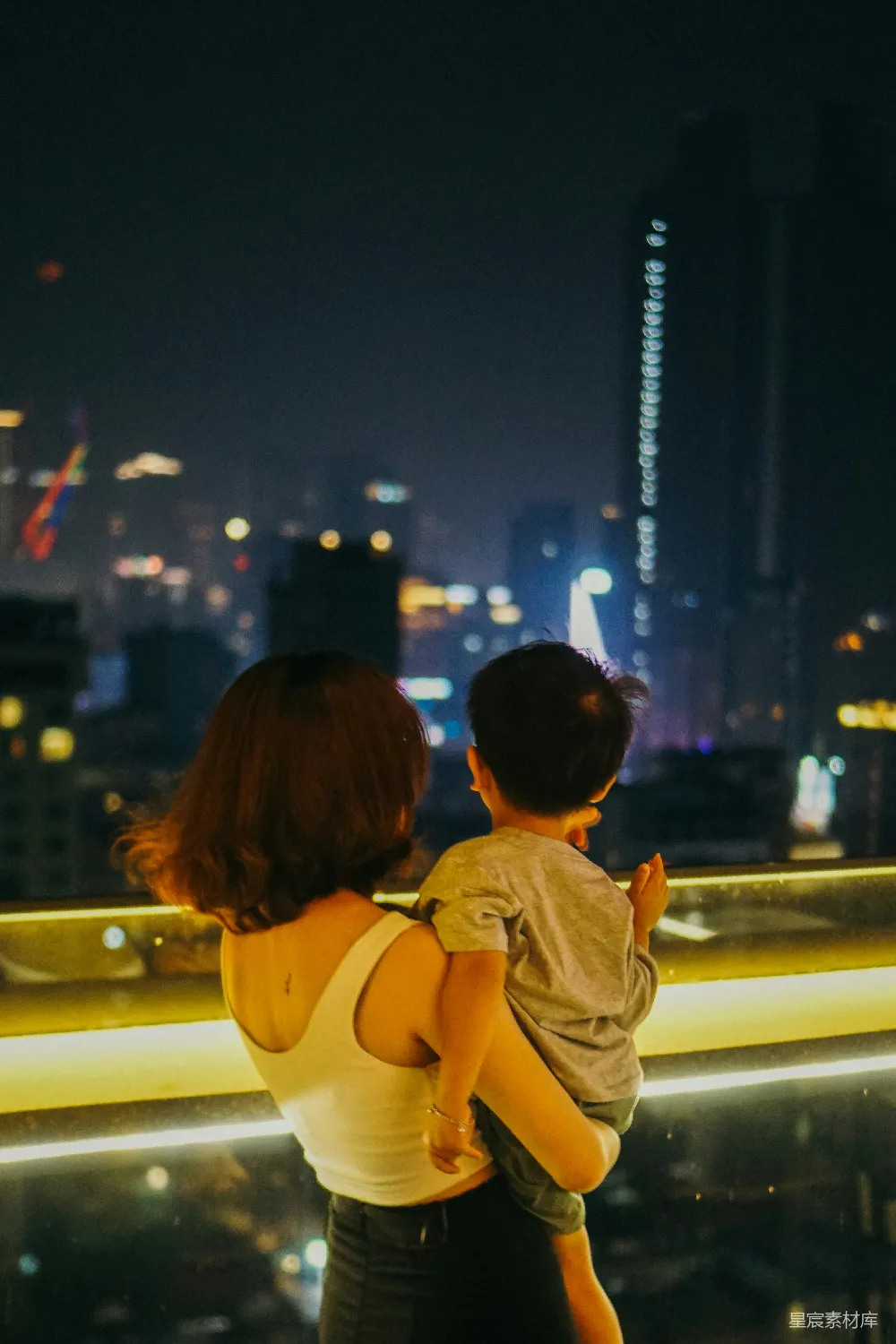 这张图片展示了一位母亲和她的儿子在夜晚的城市天际线上欣赏美丽的夜景。母亲抱着儿子，两人背对着镜头，目光投向远处的高楼大厦和灯火辉煌的城市天际线。画面充满了温馨和宁静，背景的灯光与人物的剪影形成了鲜明的对比，营造出一种温暖而宁静的氛围，适合用