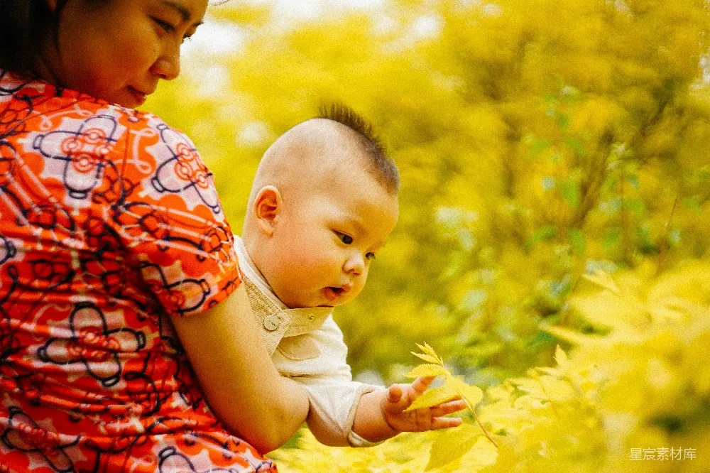 图片中，一位母亲和她的孩子在户外享受宁静的时光。母亲穿着鲜艳的红色连衣裙，抱着孩子，孩子穿着浅色的衣物，正好奇地探索着周围的树叶。背景是模糊的黄色树叶，营造出一种温馨、自然的氛围。这张照片捕捉到了母子之间深厚的情感和自然的美丽。