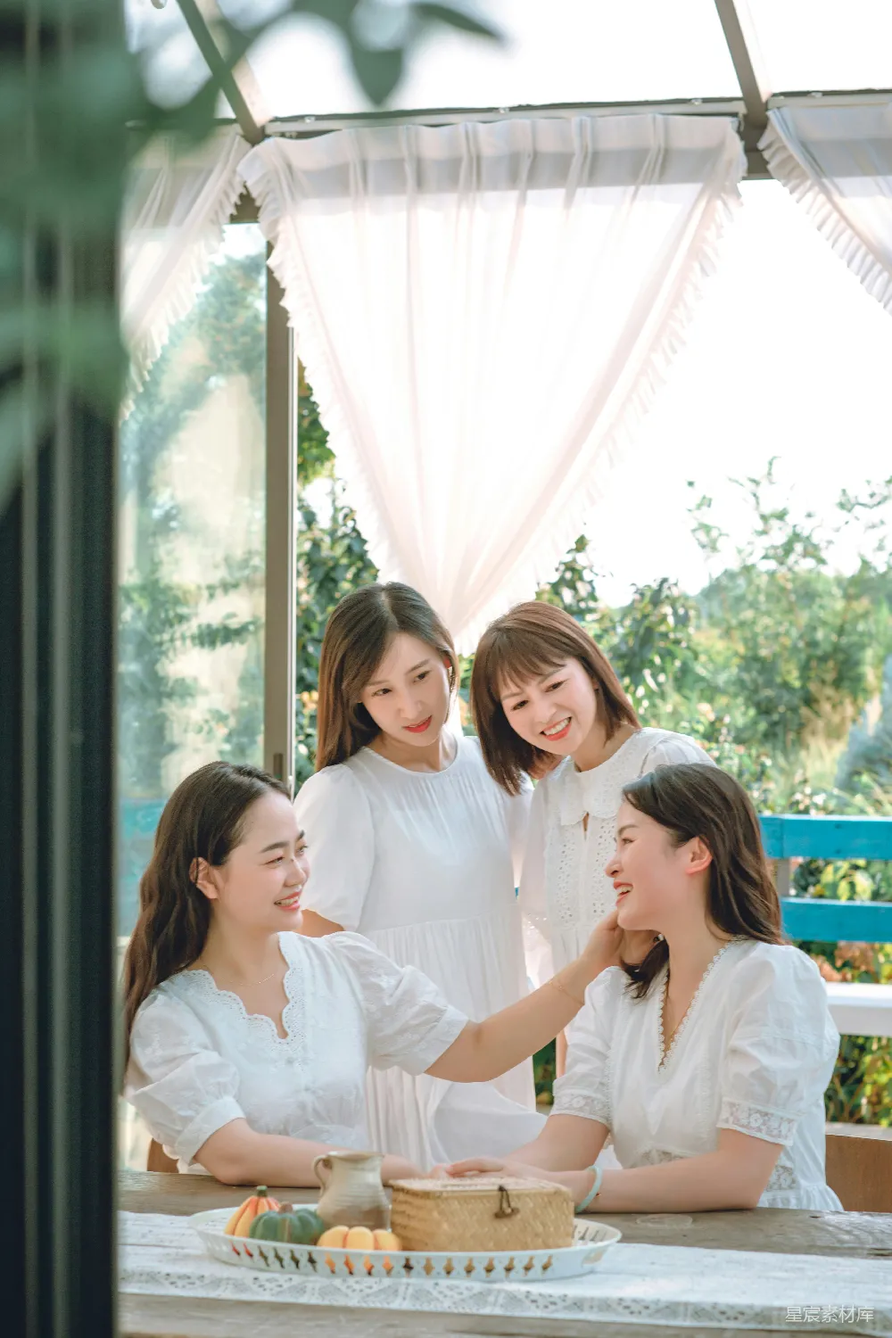 四名女性在户外的阳光下共享美好时光，她们穿着白色的衣服，围坐在一张长桌旁，桌上摆放着新鲜的水果和装饰品。背景是柔和的窗帘和自然的绿植，营造出一种轻松愉快的氛围。这幅画面传达了友谊、快乐和自然的结合。