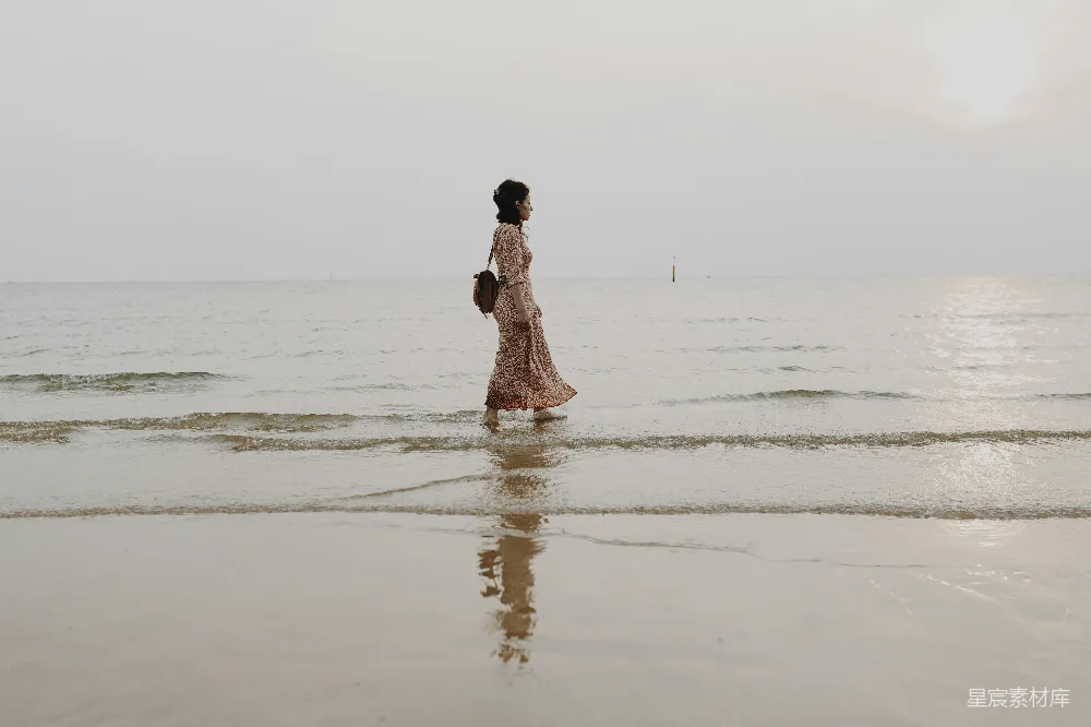 图片中，一位女性穿着碎花长裙，背对着镜头，漫步在海边。海浪轻轻拍打在她的脚边，天空有些阴沉，反射在湿润的沙滩上，营造出一种宁静而略带忧郁的氛围。整个场景充满了自然的宁静与个人的思考。