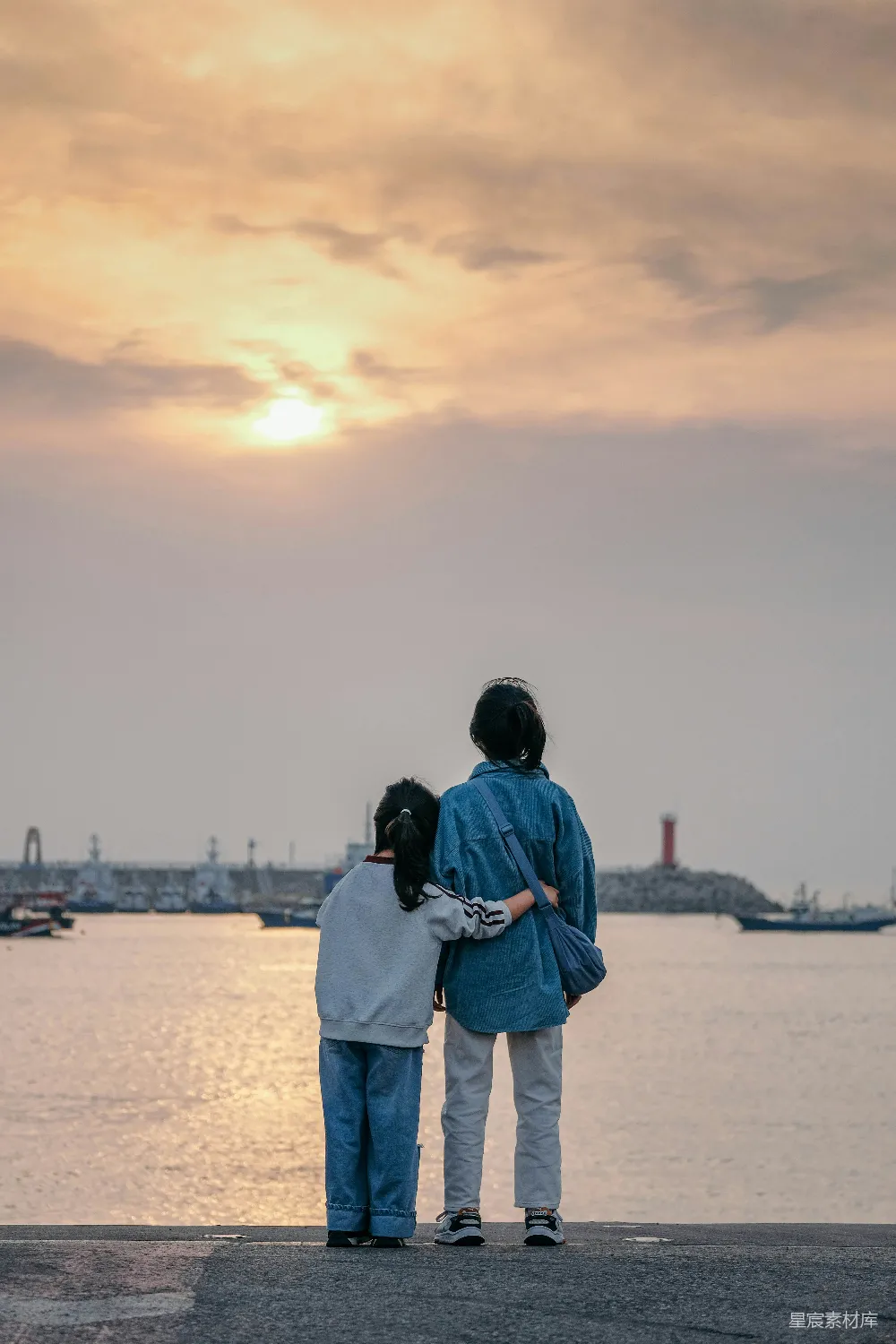 图片中，一对情侣在海边的落日余晖下相拥而立，享受着温馨的时刻。背景是宁静的海面和远处的船只，天空被染成金黄色，营造出一种宁静而浪漫的氛围。这张照片非常适合用来表现爱情的甜蜜与宁静的时光。