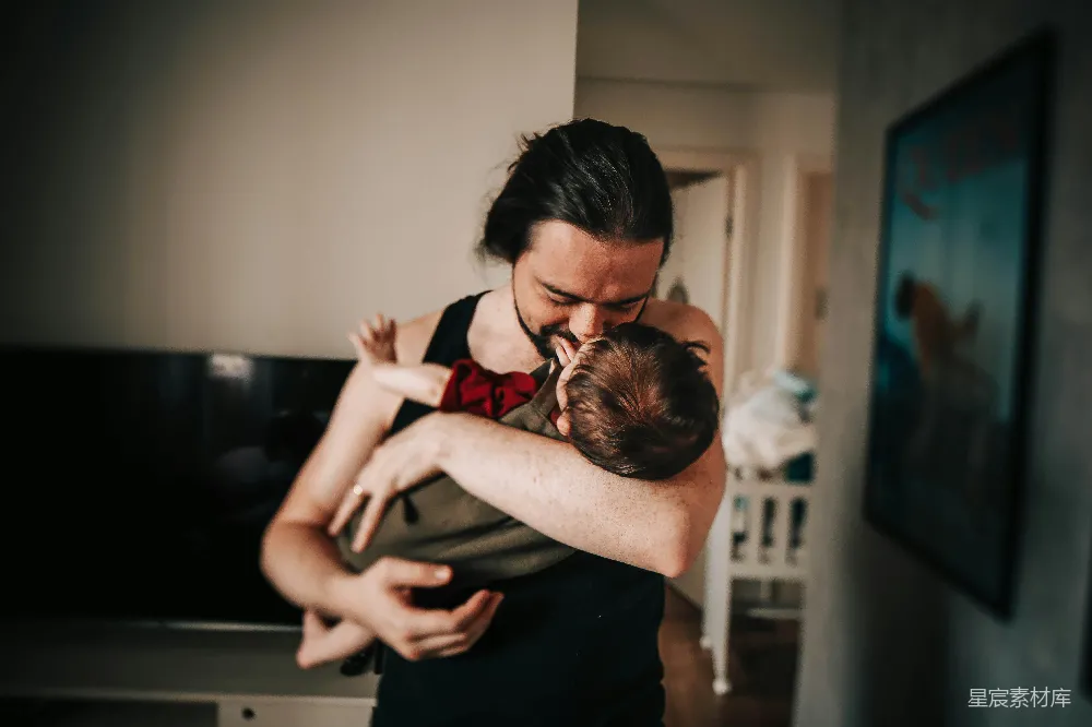 一位父亲抱着新生婴儿，深情地亲吻着孩子的额头。画面充满了温馨与爱意，背景是一个温馨的家庭环境，墙上挂着一幅画，地板上摆放着一些物品。这幅照片捕捉了家庭中最珍贵的瞬间，展现了父亲对孩子的深深关爱。