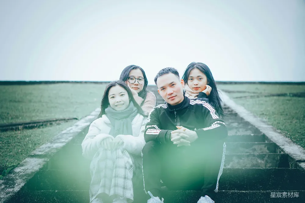 这张图片捕捉了一群年轻人在田间小径上相聚的场景，阳光透过树叶洒在他们身上，营造出温暖的氛围。他们或坐或站，轻松愉悦地交流，背景的绿色田野和开阔的天空增添了画面的宁静与自然之美。这张照片不仅记录了青春的美好时光，也展现了自然与人和谐共处的美好
