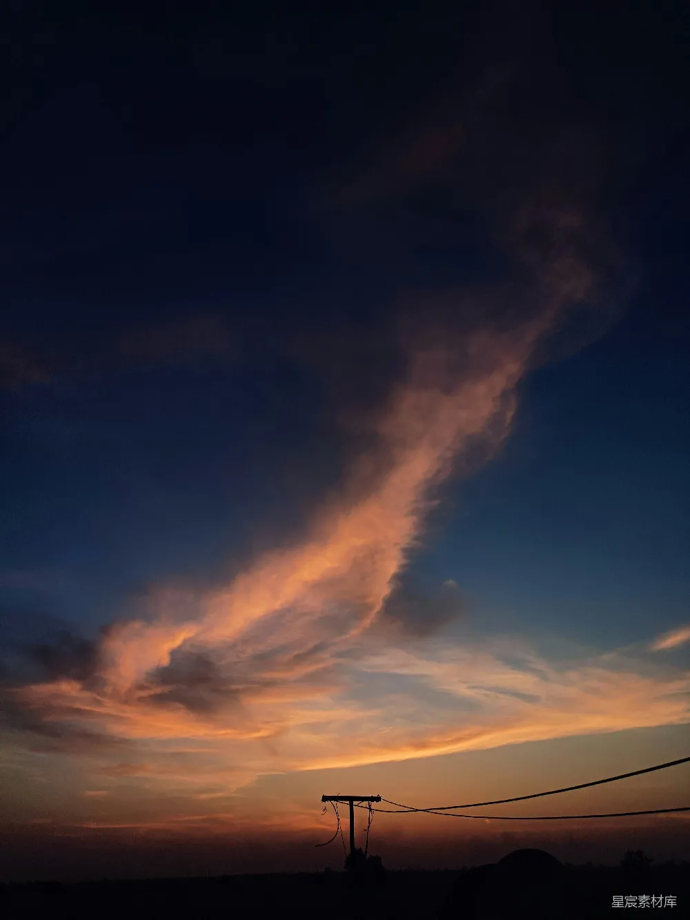 这张图片展示了黄昏时分，天空被夕阳染成橙红色，几朵云彩在空中飘动。前景中有一根电线杆的剪影，电线横跨画面，背景是连绵的山丘。整体画面宁静而美丽，展现了自然与乡村的和谐。