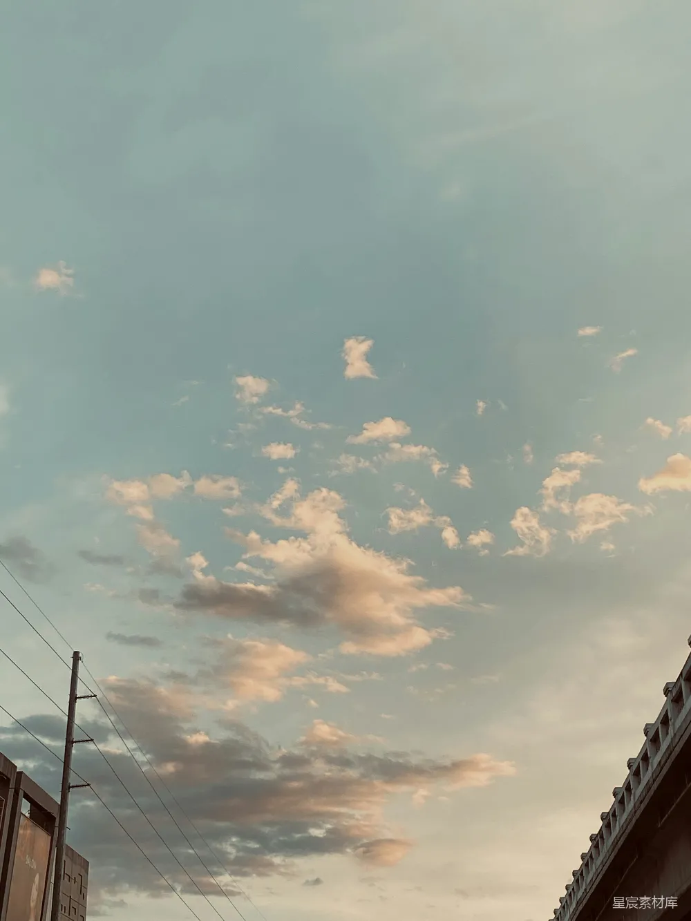 这张图片展示了黄昏时分天空中柔和的云层和远处城市的轮廓。天空中散布着几朵云彩，颜色从淡黄色到深灰色渐变，给人一种宁静而祥和的感觉。画面下方可以看到城市的建筑物轮廓，电线杆和桥梁的剪影为画面增添了层次感。整个场景显得非常和谐，适合用作自然与城