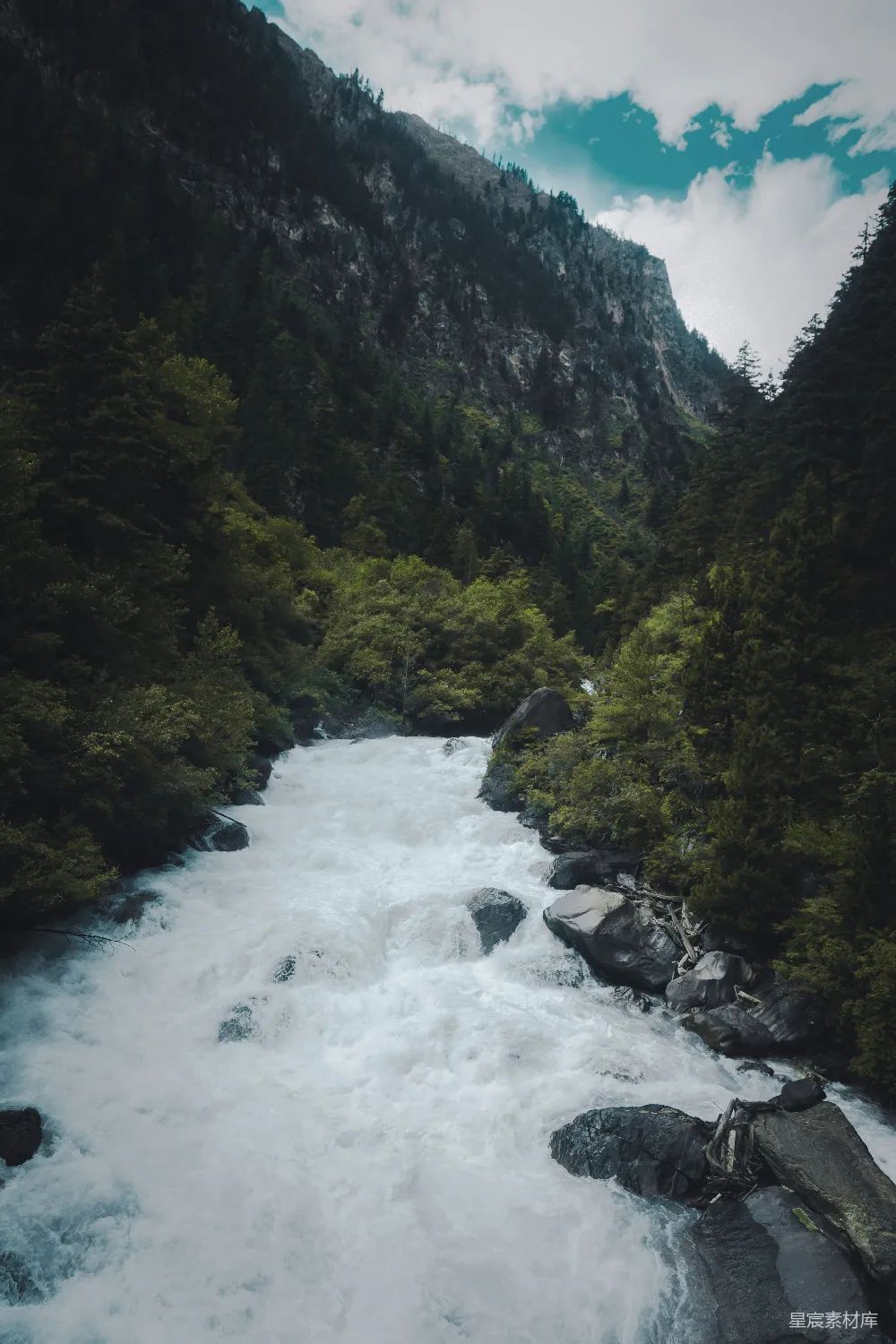 图片展示了一条湍急的河流流经山谷的场景，河水激起白色的浪花，周围是茂密的森林和陡峭的山壁。天空中飘着几朵白云，整体画面给人一种宁静而神秘的感觉，适合户外探险或自然摄影爱好者。