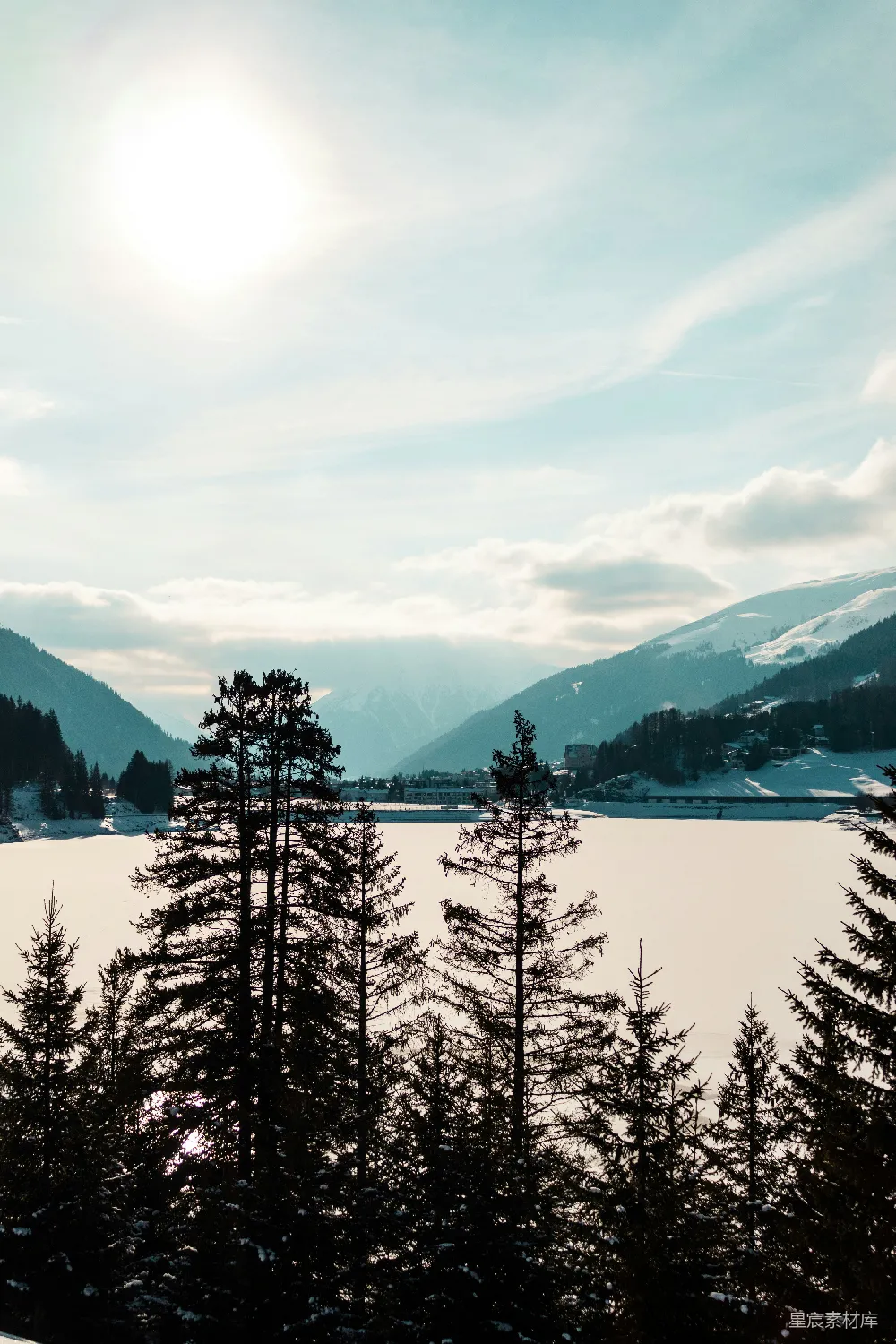 这张图片展示了一片宁静的雪地，前景是几棵高大的松树，背景是被白雪覆盖的山脉。天空中有一些云彩，阳光从云层中透出，给整个场景增添了一丝温暖。这种场景适合用作冬季旅游、自然风光和户外活动的背景图。