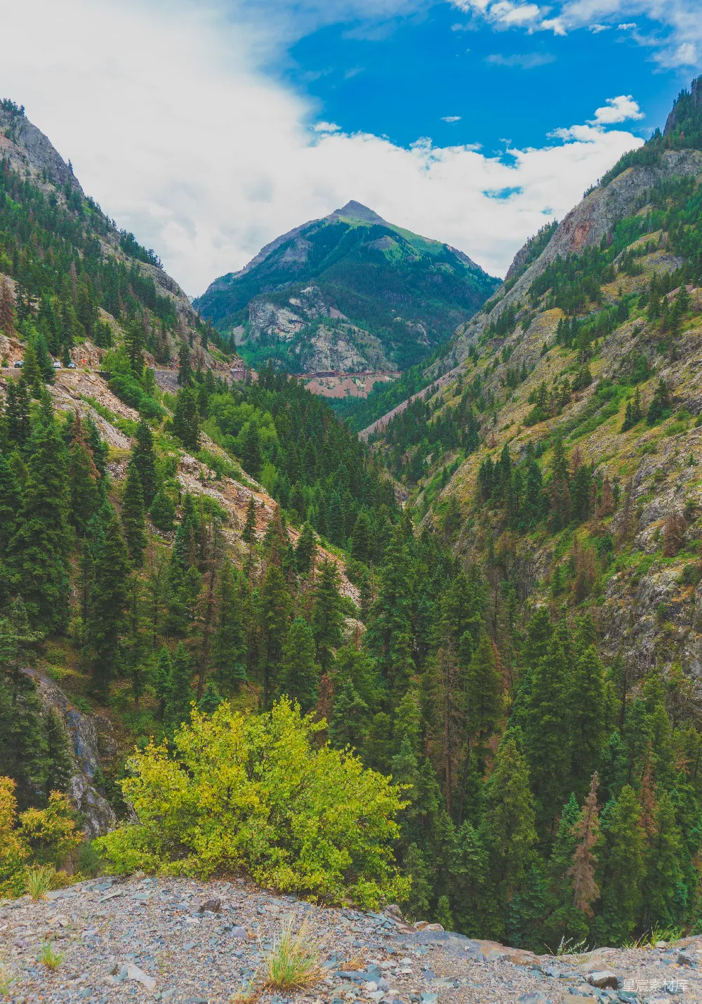 这张图片展示了一条山谷中的自然美景。两侧是高耸的山脉，山上覆盖着茂密的绿色植被。山谷中有一条蜿蜒的小路，远处可以看到一些岩石和岩石。天空中有一些白云，增加了画面的层次感。这样的景色非常适合户外旅行和摄影爱好者。