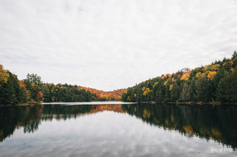 这张图片展示了秋天湖畔的宁静景色。湖面平静如镜，倒映着五彩斑斓的树叶。远处的山峦被秋色染红，天空中飘着薄薄的云层，整个画面充满了自然与和谐的美感，适合用作秋季旅游或自然摄影的素材。