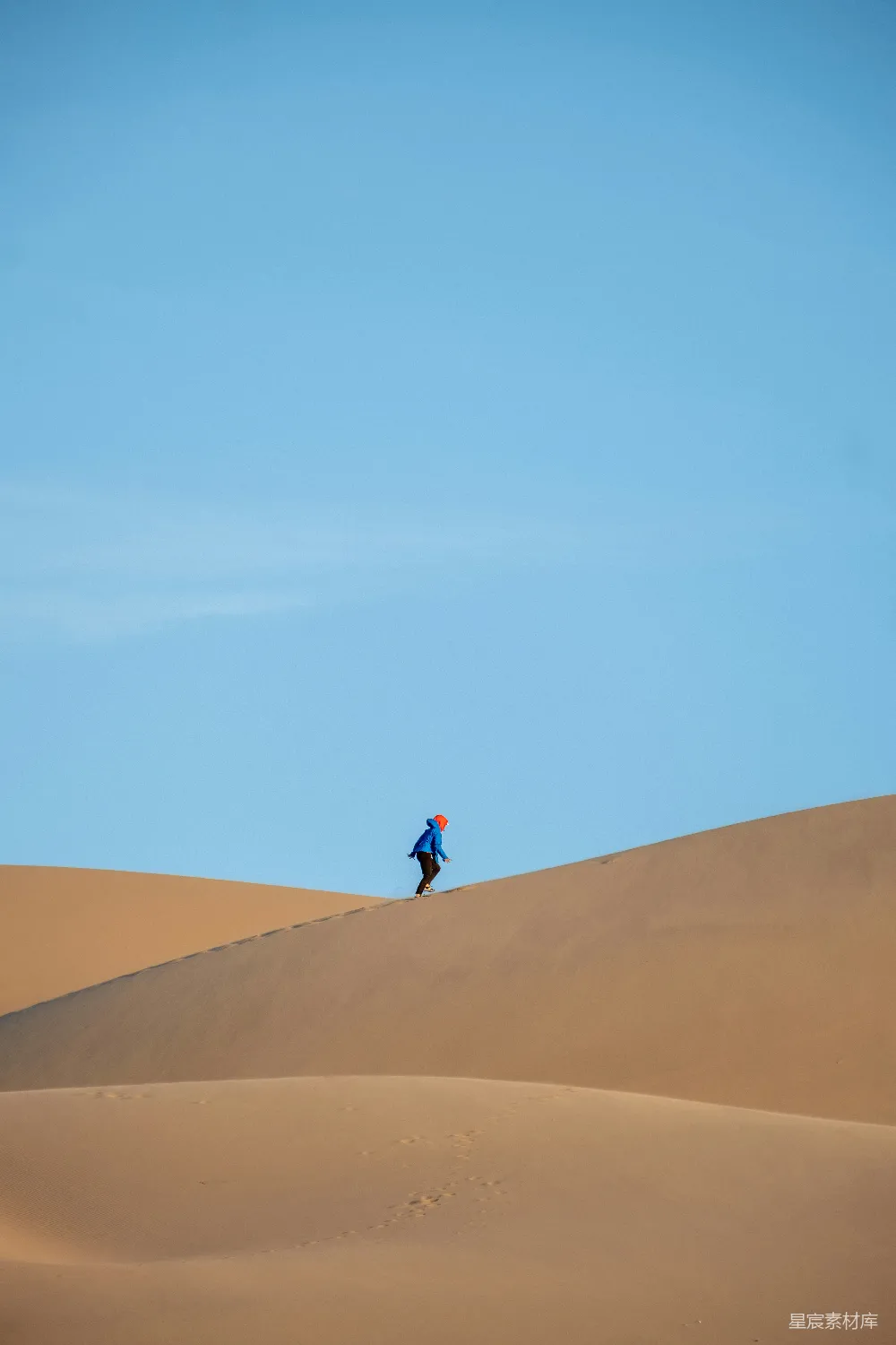 图片中，一位穿着蓝色衣服的人在广阔的沙漠中行走，背景是蓝天和沙丘。画面简洁而富有诗意，表现出一种孤独的冒险精神和对自然的敬畏。适合用于自然风光、户外探险和孤独之旅的主题。