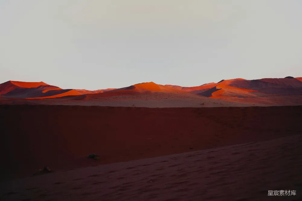 这张图片展示了沙漠中夕阳的壮丽景色。连绵起伏的沙丘被夕阳染成了红色，天空则呈现出柔和的橙色，整个场景显得宁静而壮丽。这种景象通常出现在沙漠地区，尤其是在日落时分，是旅行摄影和自然风光摄影的热门主题。