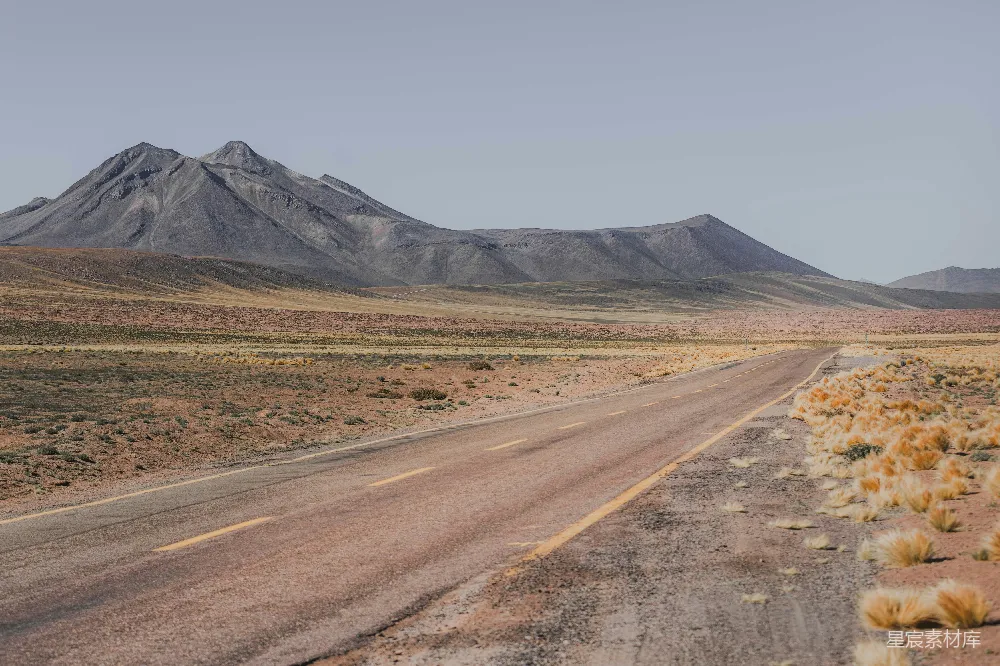 一条笔直的公路延伸至远方的山脉，周围是广阔的荒漠景观，点缀着稀疏的植被，天空晴朗，给人一种宁静而辽阔的感觉。适合用于旅行、冒险和探索自然的主题。