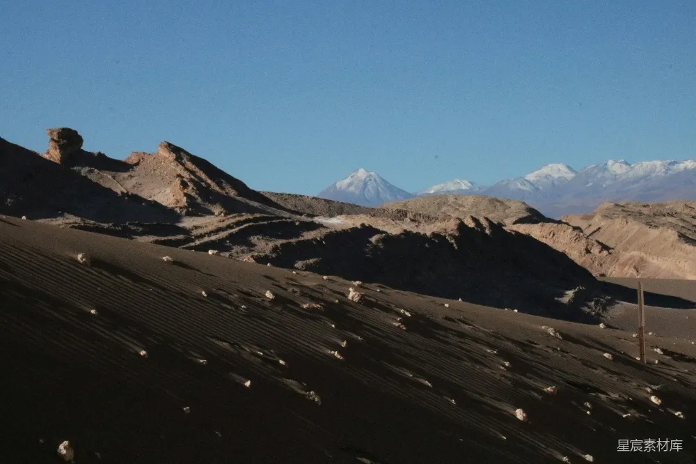 这张图片展示了一处荒凉的火山口，地面上有明显的火山灰痕迹，远处是被积雪覆盖的山峰。天空晴朗，整体色调偏冷，给人一种孤寂与神秘的感觉。适合用于自然探险、地质研究或艺术创作的背景。