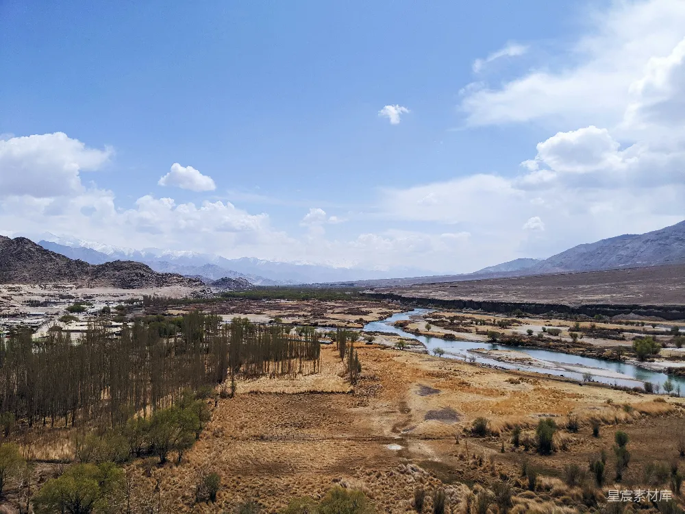 这张图片展示了一片戈壁中的河流，河流蜿蜒穿过干涸的荒地，远处是连绵的雪山。天空湛蓝，白云飘浮，给人一种宁静而辽阔的感觉。这种场景适合用于描述自然风光、荒漠探险或旅行目的地。