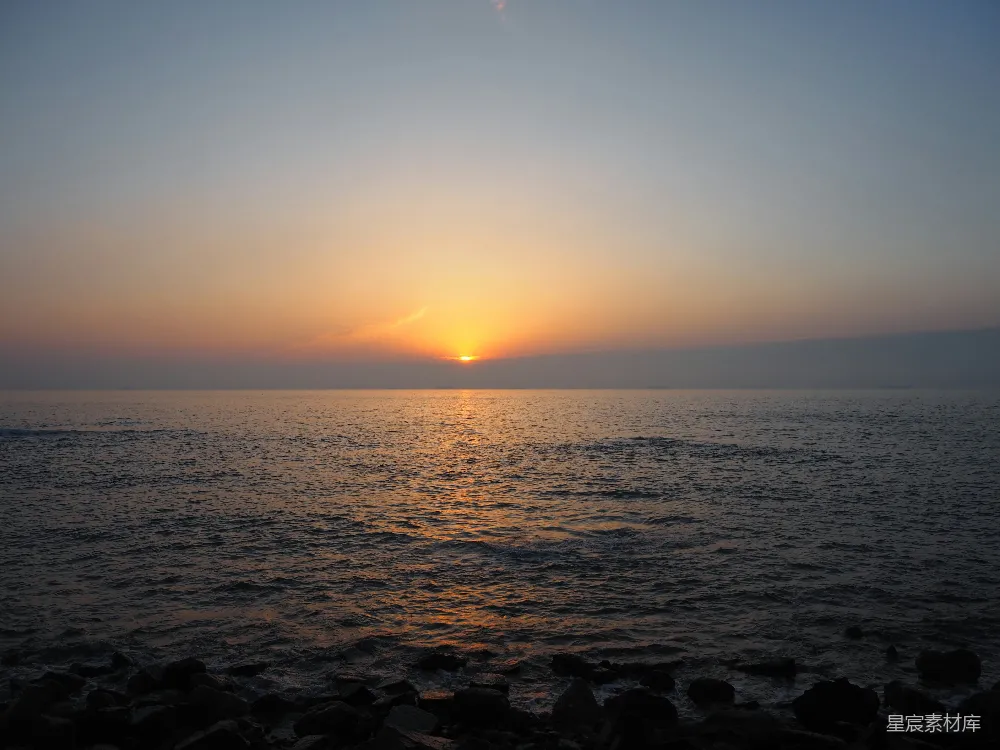 这张图片展示了海平面上的夕阳，太阳正缓缓沉入海中，金色的光芒洒在水面上，反射出温暖的色彩。天空从橙色渐变为蓝色，海面平静，远处的岩石在海浪的拍打下显得更加突出。这幅画面给人一种宁静和祥和的感觉，适合用作自然风光的壁纸或是旅行博客的封面。