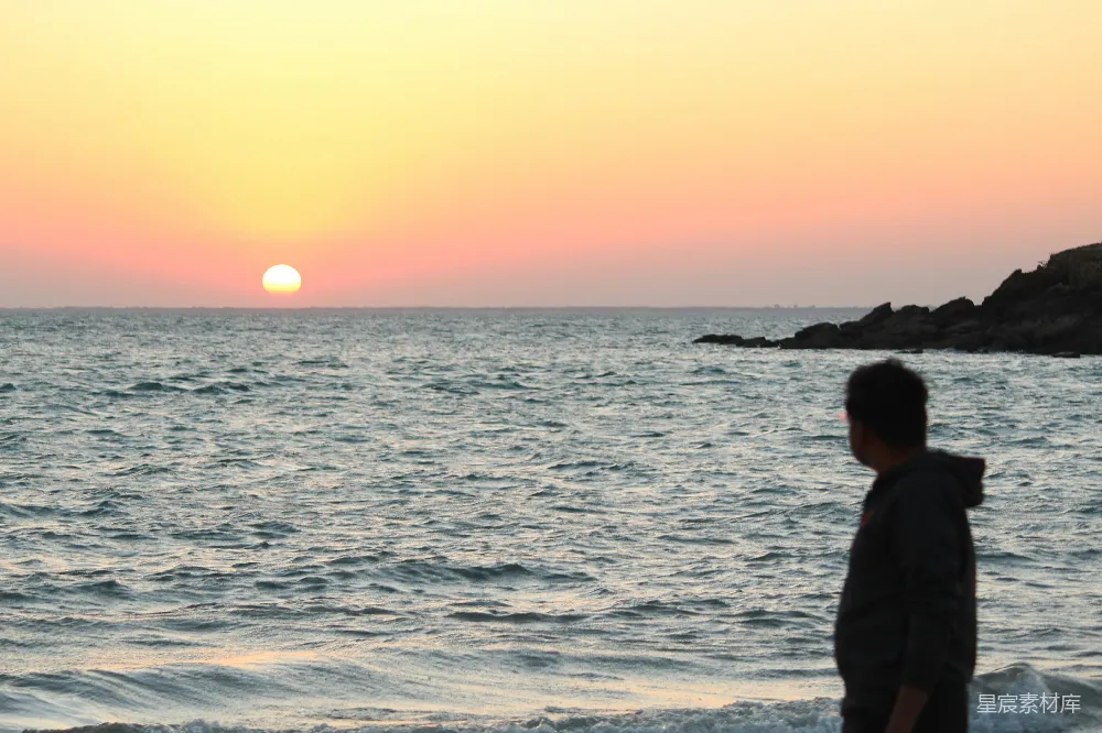 一位男子站在海边，静静地观赏夕阳西下。天空被染成了橙黄色，海面波光粼粼，远处的岩石与海浪相接，营造出一种宁静而祥和的氛围。适合用于展现自然美景、休闲时光或放松心情的主题。
