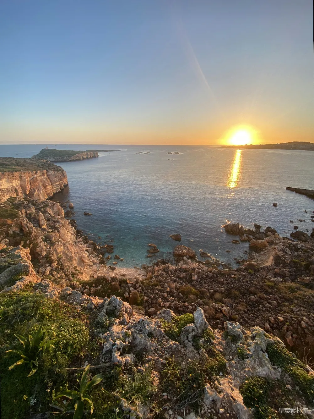 图片展示了一幅宁静的海岸线景象，夕阳的余晖洒在海面上，形成了美丽的金色反射。海岸线由岩石和少量植被组成，远处可以看到一些建筑物。整个画面给人一种宁静而美丽的自然感受，适合用来作为旅游宣传或自然风光展示。