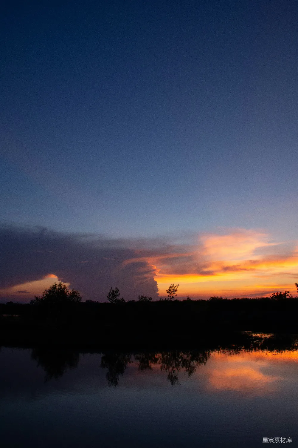 这张图片展示了宁静的湖边黄昏景色。天空被夕阳染成橙红色，云彩和湖面倒影形成美丽的对称效果。远处的树影在湖面上投下剪影，整个场景显得非常平静和美丽。适合用作自然风光或静谧场景的背景。