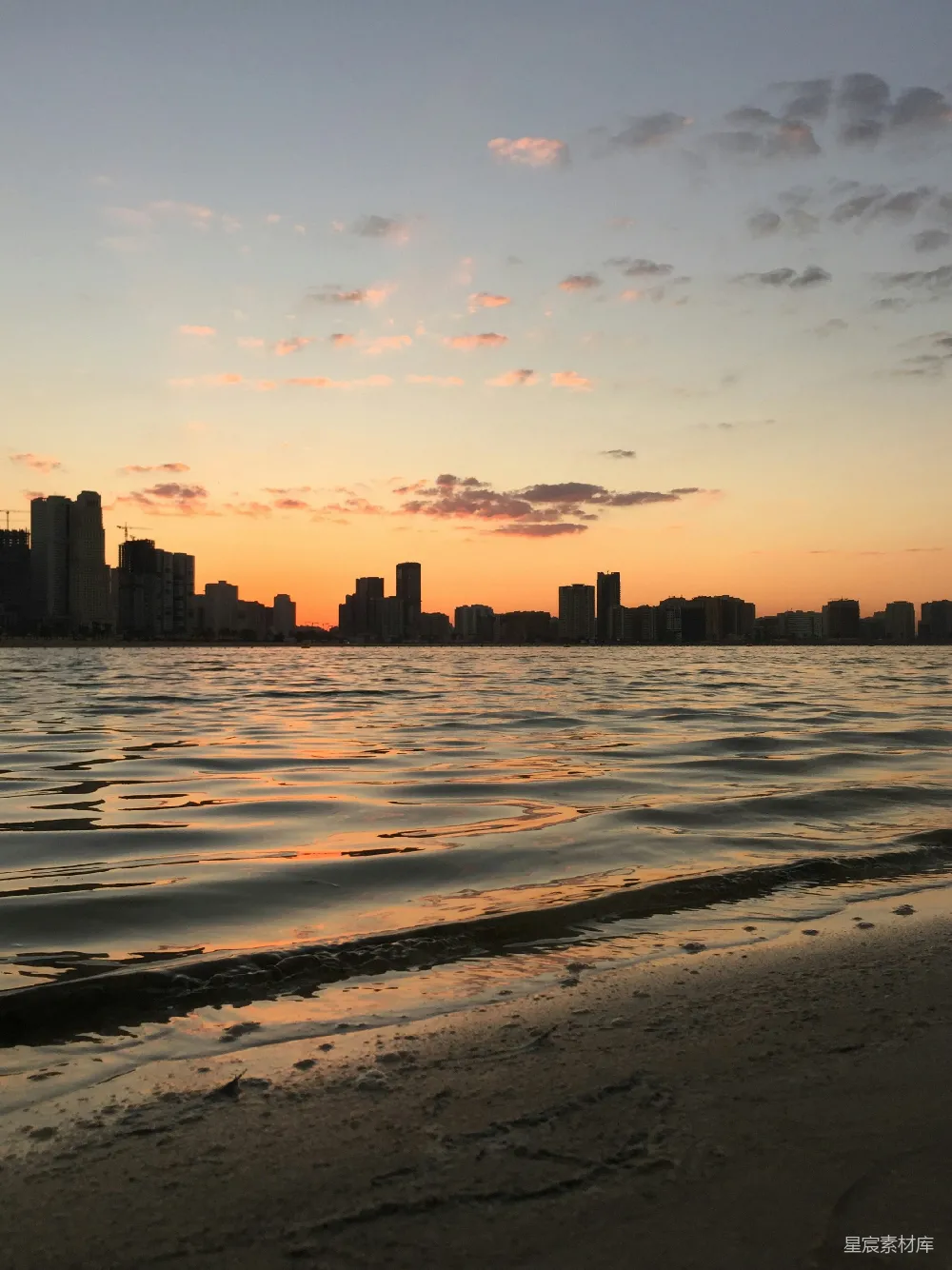 这张图片展示了黄昏时分，城市天际线的剪影与海面的宁静景象。天空被夕阳染成了橙黄色，云朵在天际飘散，海面上波光粼粼，反射出夕阳的余晖。远处的城市高楼剪影与近处的海滩形成对比，营造出一种平和而美丽的氛围。适合用于放松和旅行相关的背景。