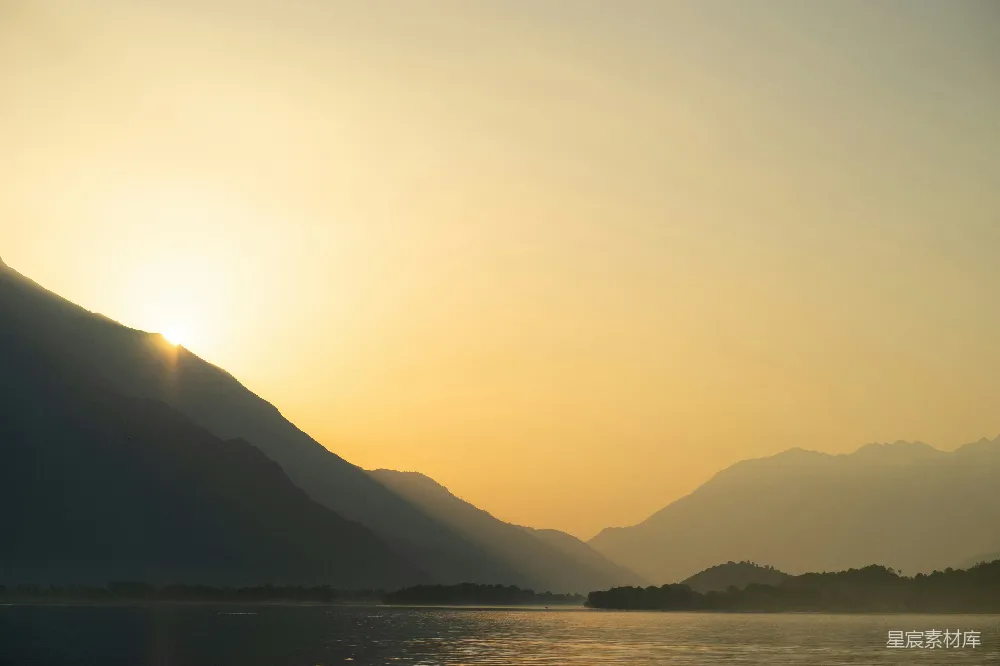 这张图片展示了一个宁静的自然景象，夕阳从山脉的一侧缓缓落下，阳光洒在水面上，形成美丽的光影效果。远处的山脉在夕阳的映衬下显得轮廓分明，整个画面给人一种宁静而祥和的感觉。适合用作自然风光、旅行、摄影等场景的背景素材。