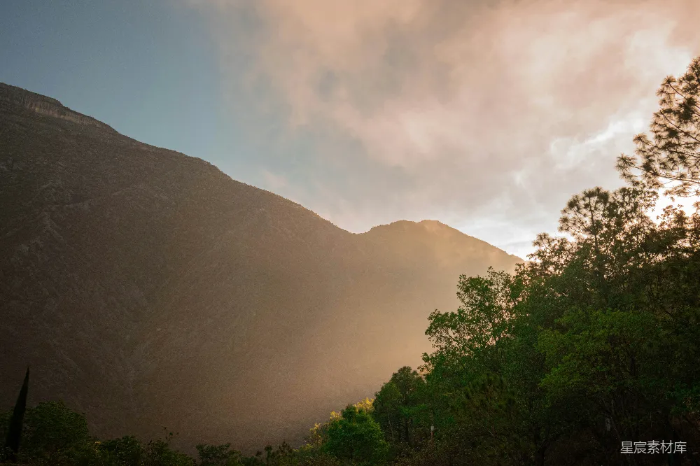 图片展示了一个夕阳西下的山谷景色，夕阳的余晖洒在山峰和树木上，形成了美丽的光影效果。天空中云彩斑斓，山峦起伏，树木郁郁葱葱，给人一种宁静而祥和的感觉。这张图片适合用于自然风光、旅游宣传或户外活动的背景素材。