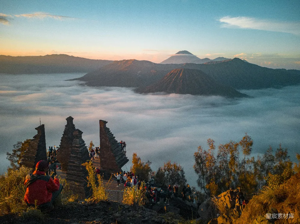 图片展示了一处日出时的婆罗摩火山景象。远处的火山在晨光中若隐若现，而近处的火山口则被一层云雾环绕，形成了壮观的云海景观。火山脚下，古迹遗址上聚集着许多游客，他们正享受着这壮丽的自然美景。整个场景充满了宁静与壮丽，是户外旅游和摄影爱好者不可多