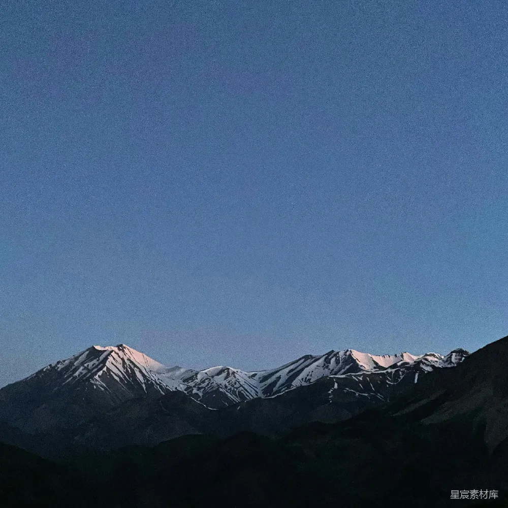 图片展示了一片黄昏时分的雪山景象，天空呈现出深邃的蓝色，山峰在夕阳的余晖下略带橙色，远处的山脉层次分明，给人一种宁静而壮丽的感觉。适合用于展现自然之美或旅行摄影。