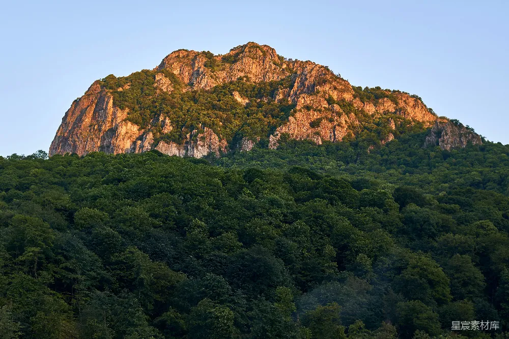 图片展示了一座被茂密森林环绕的山峦，夕阳的余晖洒在岩石峭壁上，呈现出金色的光辉，给人一种宁静而祥和的感觉。这幅画面适合用于自然风光、户外探险或宁静生活的主题背景。