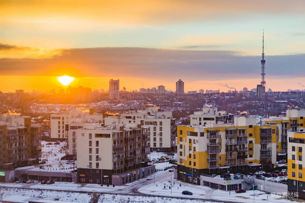 图片展示了一个冬日黄昏的城市全景，夕阳的余晖洒在城市的上空，给整个画面染上了一层温暖的橙色。远处可以看到高楼大厦和一座电视塔，近处则是低矮的住宅楼，街道上覆盖着一层薄雪，显得静谧而美丽。整个场景给人一种宁静而祥和的感觉，适合用于展示城市风光