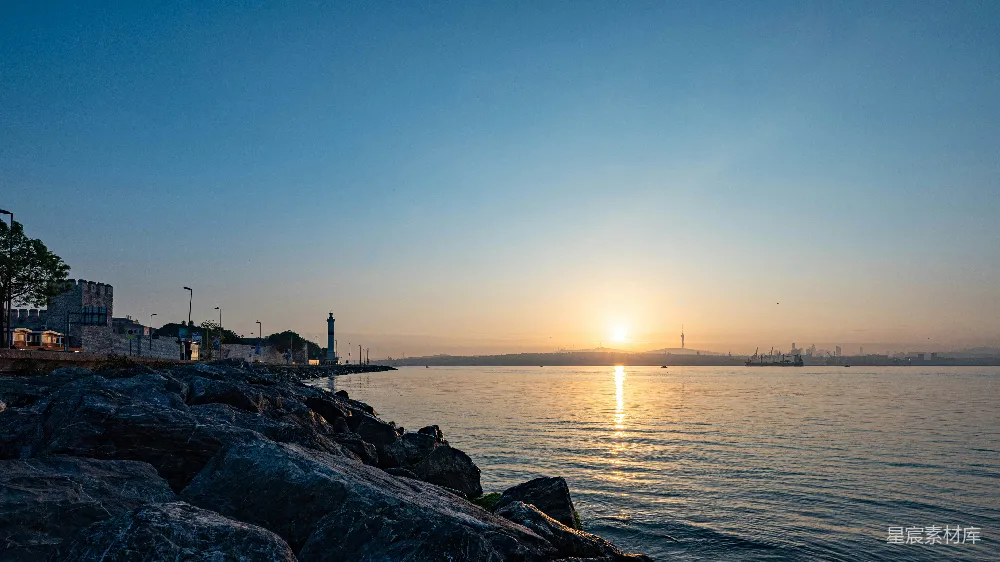 夕阳西下，金色的阳光洒在海面上，形成一道美丽的反射。岩石海岸线与远处的灯塔形成鲜明对比，远处的货船在海平线上静默地等待。整个场景宁静而美丽，适合用来表现自然之美和日落时分的宁静时刻。