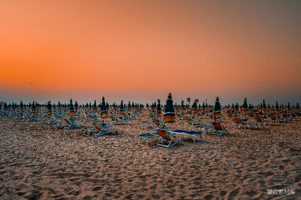这张图片展示了一片宁静的海滩，夕阳的余晖洒在沙滩上，天空被染成了温暖的橙色。沙滩上整齐排列着许多躺椅和遮阳伞，营造出一种悠闲的度假氛围。适合用于表达旅行、休闲或自然风光的主题。