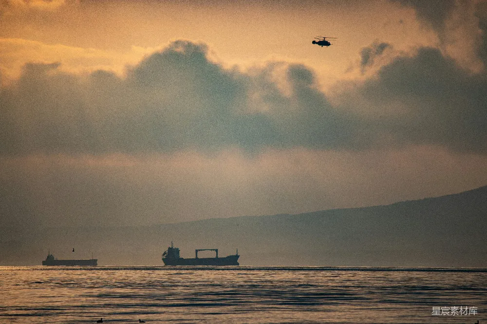 在这张图片中，夕阳的余晖洒在海面上，形成一幅宁静的海景。画面中有两艘船只静静地航行在平静的海面上，一只直升机在空中飞行，天空中云层厚重，远处隐约可见山脉的轮廓。整个场景给人一种安静、祥和的感觉，适合用作旅行、自然风光或旅游宣传图片。