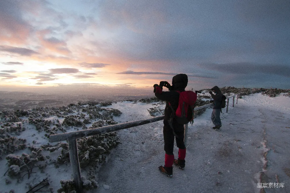 这张图片展示了两个摄影师在雪地日落时捕捉美景的场景。夕阳的余晖洒在被雪覆盖的地面上，天空被染成了柔和的橙色和蓝色。前景中的摄影师手持相机，全神贯注地拍摄着眼前这美丽的画面。远处的风景被夕阳的光辉所笼罩，营造出一种宁静而壮丽的氛围。这幅图像完
