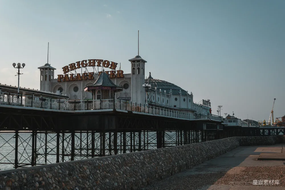 图片展示了布赖顿皇宫码头的场景。码头上的标志醒目地显示着'BRIGHTON PALACE PIER'，背景是蓝天和远处的工业起重机。这座建筑以其独特的设计和标志性的外观吸引着游客。